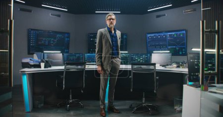 Photo for Mature big data scientist in business suit puts on glasses, stands in monitoring room and looks at camera. Multiple big screens on the wall with displayed live analysis feed at background. Dolly shot. - Royalty Free Image