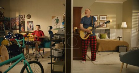 Photo for Young boy practices musician skills uses drum kit. Elderly man sings and plays on electric guitar with neighbour. Thin walls and low level of sound insulation. Neighbors play music together. - Royalty Free Image