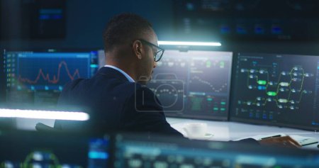 Téléchargez les photos : Ingénieur logiciel afro-américain assis devant l'ordinateur et les grands écrans numériques avec serveur de données et réseau blockchain, travaille dans la salle de contrôle de surveillance moderne avec des graphiques d'analyse en temps réel. - en image libre de droit
