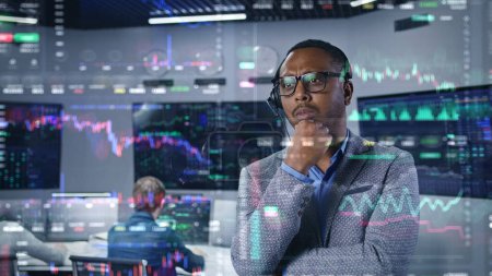 African American trader, businessman works in modern broker agency office. 3D real-time stocks and cryptocurrency charts on glass wall. VFX animation. Computers and big digital screens on background.