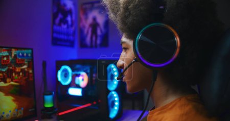 Photo for Happy gamer in headphones wins a round in multiplayer game on home personal computer. African American teenager plays third person shooter. Online video game live streaming or cybersport competition. - Royalty Free Image