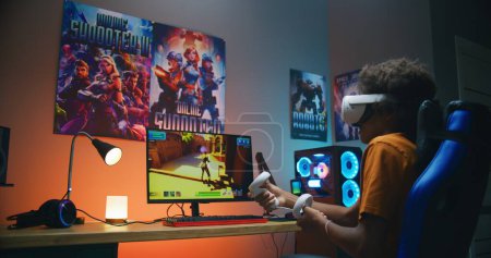 Téléchargez les photos : Afro-Américain adolescent gamer dans VR casque joue tireur 3D à la troisième personne virtuelle sur ordinateur personnel à l'aide de contrôleurs sans fil. Jeu vidéo en ligne en streaming ou tournoi d'e-sport. Jeu à la maison. - en image libre de droit