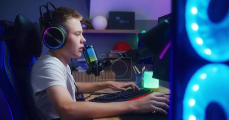 Photo for Young player in headphones talks with teammates using professional microphone. Schoolboy plays PvP multiplayer game on computer at home. Online video game live streaming or cybersport championship. - Royalty Free Image