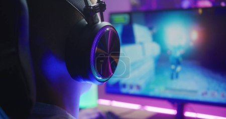 Young boy in headphones plays third person shooter. Gamer enjoys online video game on home personal computer. Desk illuminated by RGB LED strip light. Concept of gaming at home. Back view. Close up.