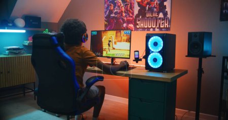 Photo for African American young gamer in headphones plays online third-person 3D shooter on modern PC in stylish neon room. Video game live streaming or cybersport competition. Gaming at home. - Royalty Free Image