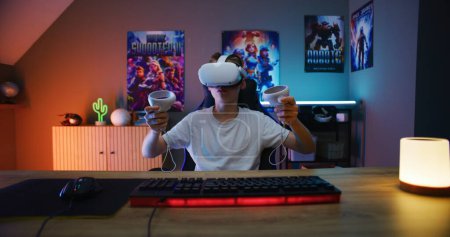 Photo for Young gamer in VR headset plays virtual online video game using wireless controllers. Boy enjoys eSports tournament. Room with warm neon LED lights. Gaming at home. POV from PC screen perspective. - Royalty Free Image