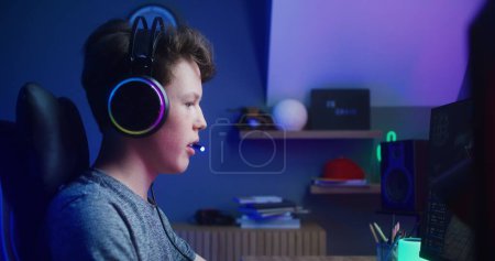 Photo for Concentrated boy in headphones plays in multiplayer game on PC at home. Gamer talks with teammates using microphone. Online video game live streaming or esports tournament. Side view. - Royalty Free Image