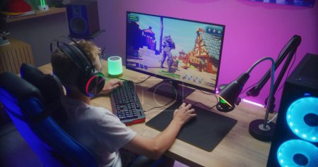 Photo for Young gamer plays third person shooter on personal computer. Schoolboy in headphones enjoys online video game at home. Desk illuminated by RGB LED strip light. Concept of gaming at home. Back view. - Royalty Free Image