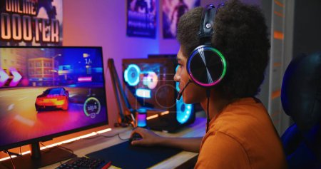 Photo for African American guy, teenage gamer in headphones plays in online 3D race game on modern powerful PC in neon cozy room. Video game live streaming or professional eSports tournament. Gaming at home. - Royalty Free Image