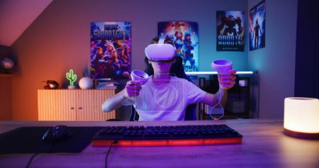 Téléchargez les photos : Jeune joueur en VR casque joue à la réalité virtuelle jeu vidéo en ligne à l'aide de contrôleurs sans fil. Garçon s'amuser dans les loisirs dans la chambre néon élégant. Jeu à la maison. Vue de la perspective de l'écran PC. - en image libre de droit