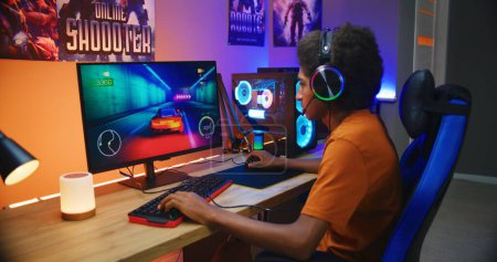African American guy, teenage gamer in headphones plays in online 3D race game on modern powerful PC in neon cozy room. Video game live streaming or professional eSports tournament. Gaming at home.