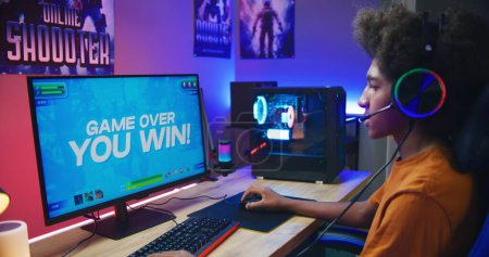 African American gamer plays in third person shooter on PC at home. Computer monitor with displayed online video game live stream or cybersport tournament. Teenager spending leisure time.