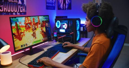 Jugador afroamericano juega en tercera persona shooter en PC en casa. Monitor de computadora con visualización de video juegos en línea en vivo o torneo de ciberdeporte. Adolescente pasar tiempo libre.