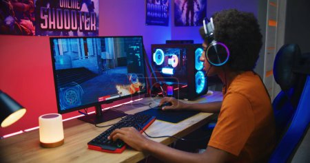 Photo for African American teenage gamer in headphones plays and loses round in online first-person 3D shooter on modern PC in neon room. PvP video game live streaming or eSports tournament. Gaming at home. - Royalty Free Image
