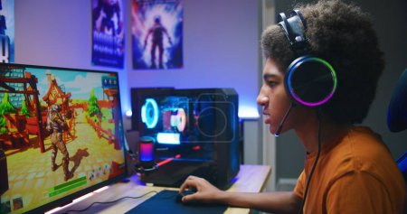 Photo for Professional eSports African American young gamer puts on headphones and plays online third-person 3D shooter on modern PC in stylish neon room. Video game live streaming or cybersport competition. - Royalty Free Image