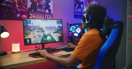 Photo for African American teenager in headphones talks with teammates, plays online tank battling 3D game on PC in neon room. Video game live streaming or professional cybersport championship. Gaming at home. - Royalty Free Image