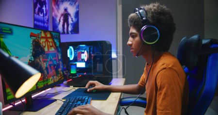Photo for Professional eSports African American young gamer puts on headphones and plays online third-person 3D shooter on modern PC in stylish neon room. Video game live streaming or cybersport competition. - Royalty Free Image