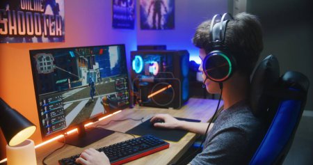 Photo for Young gamer plays in first person shooter on PC at home. Computer monitor with displayed online video game live stream or cybersport championship. Desk illuminated by RGB LED strip light. - Royalty Free Image