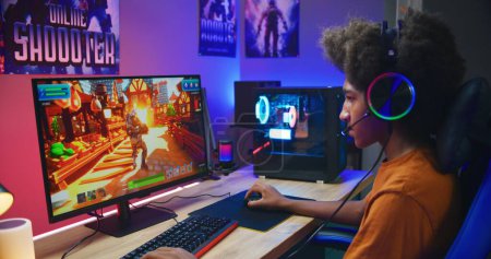 Photo for African American gamer plays in third person shooter on PC at home. Computer monitor with displayed online video game live stream or cybersport tournament. Teenager spending leisure time. - Royalty Free Image