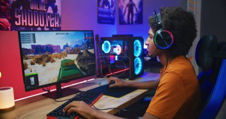 Photo for African American teenager in headphones talks with teammates, plays online tank battling 3D game on PC in neon room. Video game live streaming or professional cybersport championship. Gaming at home. - Royalty Free Image