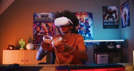 Photo for Teenage gamer in VR headset plays virtual online video game using wireless controllers. African American guy having fun in leisure time in his room. Gaming at home. View from PC screen perspective. - Royalty Free Image