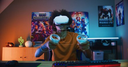 Téléchargez les photos : Teenage gamer in VR casque joue jeu vidéo virtuel en ligne à l'aide de contrôleurs sans fil. Afro-Américain s'amuser dans ses loisirs dans sa chambre. Jeu à la maison. Vue de la perspective de l'écran PC. - en image libre de droit