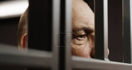 Téléchargez les photos : Visage de près d'un prisonnier âgé déprimé se tenant derrière des barreaux en métal dans une cellule de prison. Un criminel ou un meurtrier purge une peine d'emprisonnement pour crime. Détenu en prison ou en centre de détention. Système judiciaire. - en image libre de droit