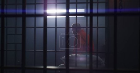 Anciano criminal en uniforme naranja se sienta en la celda, se levanta y mira a la ventana con barras. Recluso culpable en un centro de detención o correccional. Prisionero cumple pena de prisión.