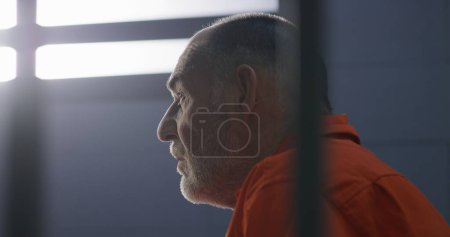 Foto de Anciano criminal con uniforme naranja se sienta en la cama de la prisión y sueña con la libertad. El prisionero cumple condena de prisión en la celda. Recluso sombrío en centro de detención o correccional. - Imagen libre de derechos