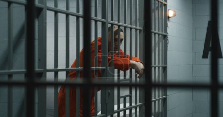 Foto de Prisionero de edad avanzada en uniforme naranja estira los dedos, se apoya en barras de metal. Criminal cumple pena de prisión por crimen en celda de prisión. Recluso cansado tras las rejas en la cárcel o centro de detención. - Imagen libre de derechos