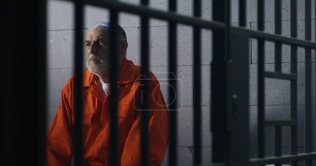 Foto de Anciano criminal en uniforme naranja se sienta en la cama de la prisión y piensa en la libertad. El prisionero cumple condena de prisión en la celda. Recluso culpable en centro de detención o correccional. - Imagen libre de derechos