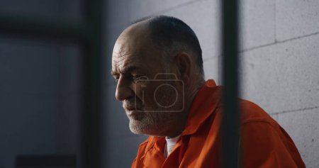 Photo for Elderly criminal in orange uniform sits on prison bed and dreams about freedom. Prisoner serves imprisonment term in jail cell. Gloomy inmate in detention center or correctional facility. - Royalty Free Image