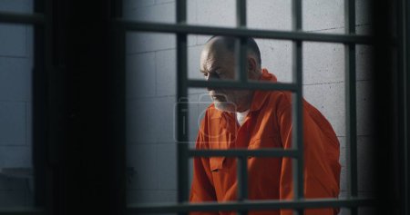 Foto de Anciano criminal en uniforme naranja se sienta en la cama de la prisión y piensa en la libertad. El prisionero cumple condena de prisión en la celda. Recluso culpable en centro de detención o correccional. - Imagen libre de derechos