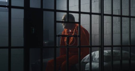 Elderly criminal in orange uniform sits on jail bed and talks with cellmate. Depressed prisoners serve imprisonment term in prison cell. Guilty inmates in detention center or correctional facility.