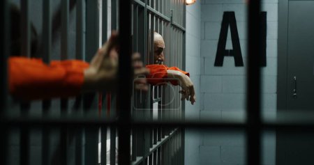 Foto de Los prisioneros con uniformes naranjas se apoyan en barras de metal en las celdas de la prisión. Los criminales culpables cumplen penas de prisión por crímenes en la cárcel, centro de detención. El preso mayor habla agresivamente. Vista desde la celda - Imagen libre de derechos