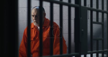 Téléchargez les photos : Un criminel âgé en uniforme orange est assis sur un lit de prison et pense à la liberté. Le prisonnier purge une peine d'emprisonnement en cellule. Détenu coupable dans un centre de détention ou un établissement correctionnel. - en image libre de droit