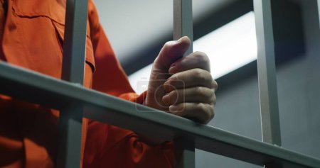 Großaufnahme eines Gefangenen in orangefarbener Uniform mit Metallstangen, der in der Gefängniszelle steht. Schuldiger Verbrecher oder Mörder verbüßt eine Gefängnisstrafe für ein Verbrechen. Häftling im Gefängnis oder in der Haftanstalt. Justizsystem.