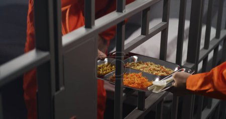 Photo for Elderly prisoner in orange uniform sits in prison cell. Prison guard gives him tray of food through metal bars. Guilty criminal serves imprisonment term for crime. Inmate in jail or detention center. - Royalty Free Image
