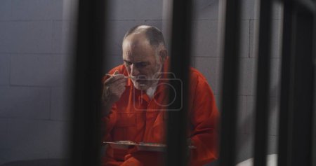 Photo for Elderly prisoner in orange uniform sits in prison cell. Prison guard gives him tray of food through metal bars. Guilty criminal serves imprisonment term for crime. Inmate in jail or detention center. - Royalty Free Image