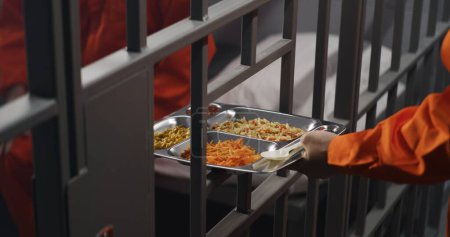 Foto de Un preso mayor con uniforme naranja se sienta en la celda de la prisión. El guardia de la prisión le da bandeja de comida a través de barras de metal. El criminal culpable cumple pena de prisión por crimen. Recluso en la cárcel o centro de detención. - Imagen libre de derechos