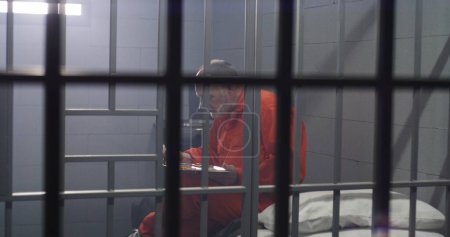 Foto de Un preso mayor con uniforme naranja se sienta en la celda de la prisión, come comida. El criminal cumple pena de prisión por crimen. Condiciones en la cárcel o correccional. Vista desde la celda de la cárcel a través de barras de metal. - Imagen libre de derechos