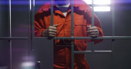 Photo for Elderly prisoner in orange uniform holds metal bars, stands in prison cell. Guilty criminal or thief serves imprisonment term for crime. Inmate in jail, detention center or correctional facility. - Royalty Free Image