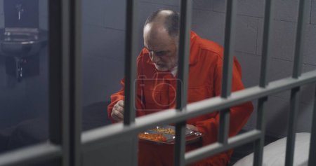 Foto de Un preso mayor con uniforme naranja se sienta en la celda de la prisión, come comida. El criminal cumple pena de prisión por crimen. Condiciones en la cárcel o correccional. Vista desde la celda de la cárcel a través de barras de metal. - Imagen libre de derechos