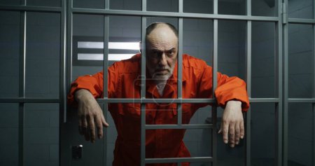 Téléchargez les photos : Prisonnier âgé en uniforme orange tient la main sur des barres métalliques, regarde la caméra. Le criminel purge une peine d'emprisonnement en cellule. Le détenu se tient derrière les barreaux en prison ou dans un centre de détention. Portrait. - en image libre de droit