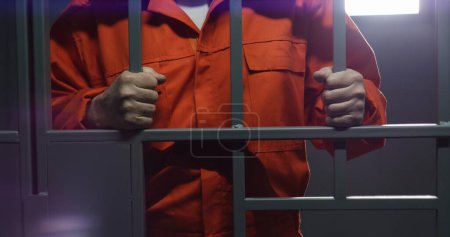 Téléchargez les photos : Un prisonnier âgé en uniforme orange tient des barres métalliques, se tient dans une cellule de prison. Un criminel ou un voleur coupable purge une peine d'emprisonnement pour crime. Détenu en prison, centre de détention ou établissement correctionnel. - en image libre de droit