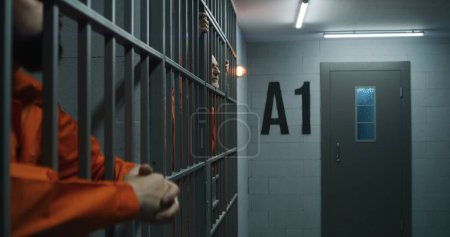 Téléchargez les photos : Prisonnier âgé en uniforme orange étire les doigts, s'appuie sur des barres métalliques. Le criminel purge une peine d'emprisonnement pour crime en cellule. Un détenu fatigué se tient derrière les barreaux en prison ou dans un centre de détention. - en image libre de droit