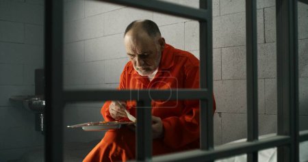Téléchargez les photos : Prisonnier âgé, détenu en uniforme orange mange de la nourriture assise dans une cellule de prison. Le criminel purge une peine d'emprisonnement pour crime. Emprisonnement, centre de détention ou établissement correctionnel. Tirer à travers les barres métalliques. - en image libre de droit