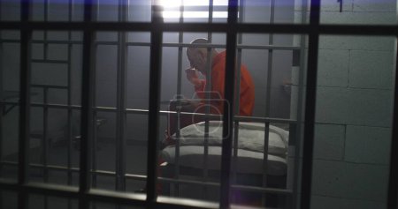 Foto de Un preso mayor con uniforme naranja se sienta en la celda de la prisión, come comida. El criminal cumple pena de prisión por crimen. Condiciones en la cárcel o correccional. Disparos desde la celda a través de barras de metal. - Imagen libre de derechos
