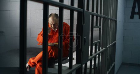 Téléchargez les photos : Prisonnier âgé, détenu en uniforme orange mange de la nourriture assise dans une cellule de prison. Le criminel purge une peine d'emprisonnement pour crime. Emprisonnement, centre de détention ou établissement correctionnel. Tirer à travers les barres métalliques. - en image libre de droit