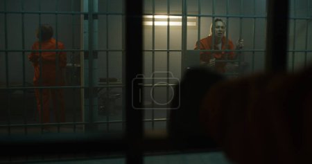 Foto de Una prisionera loca con uniforme naranja le grita a la reclusa vecina, salta a las barras de metal en la celda de la prisión. Las mujeres cumplen penas de prisión en prisión. Criminales en correccional. Sistema de justicia. - Imagen libre de derechos